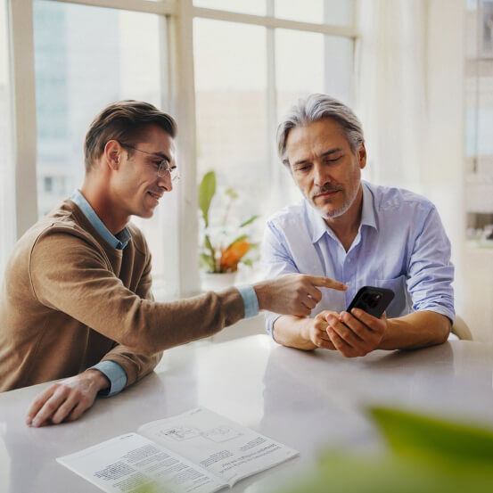 Two men in a bright office setting look together at a smartphone screen. One gestures toward the phone while an open book lies on the table nearby.